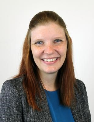 This is a photo of Amanda Blocker who's a white woman with freckles, blue eyes, and shoulder length red hair. She's wearing a gray blazer and is smiling at the camera.